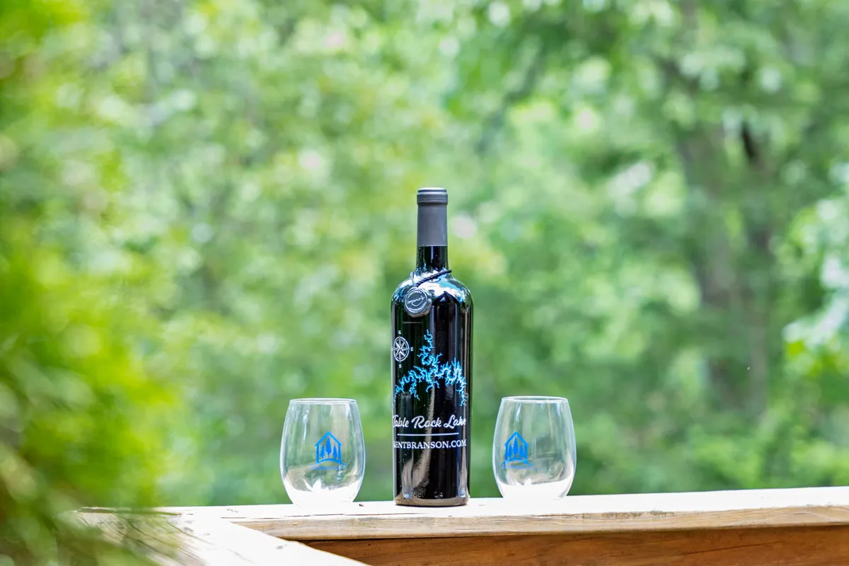 An outdoor scene focusing on a wine bottle and two glasses set up, indicating an intimate moment of wine tasting or relaxation. The background suggests a tranquil, natural environment, enhancing the mood of romantic outdoor gatherings.
