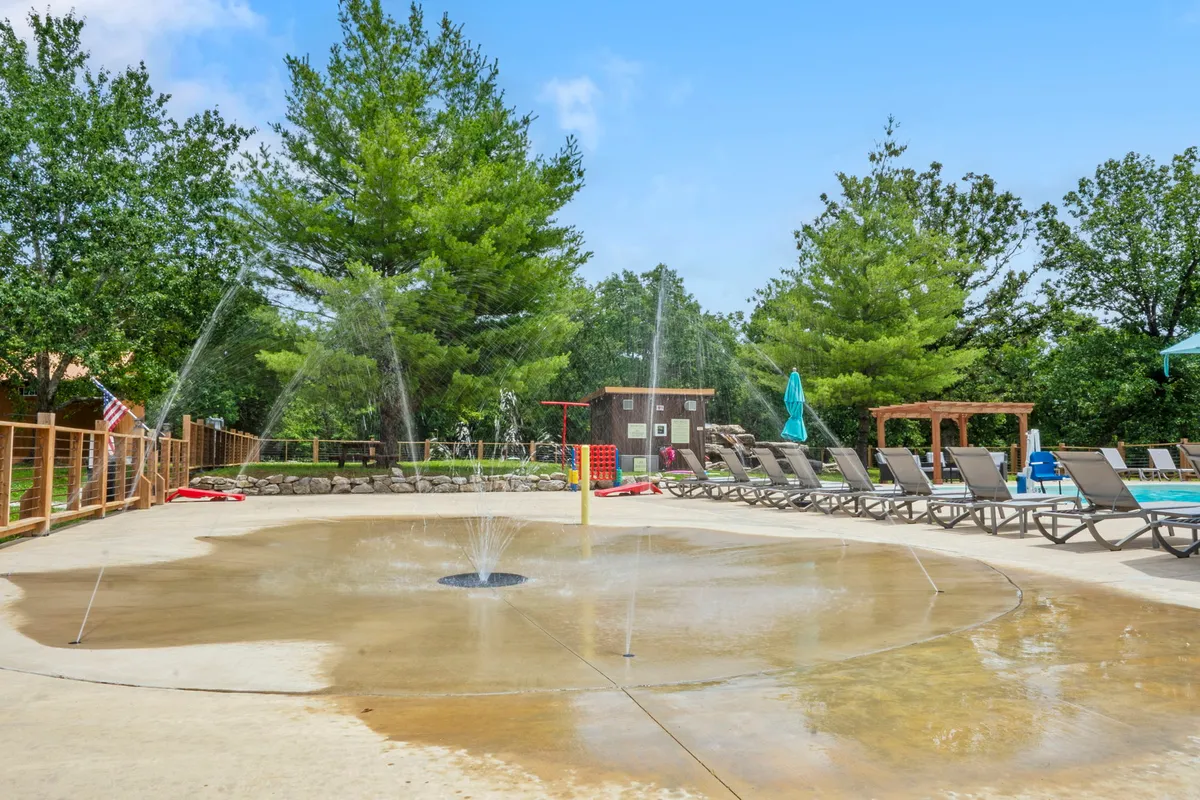 An outdoor splash pad with children playing in the water, surrounded by lounge chairs and lush greenery. Water jets and splashing features create an energetic and joyful atmosphere perfect for families. The image conveys a sense of community fun and relaxation for kids and adults alike.