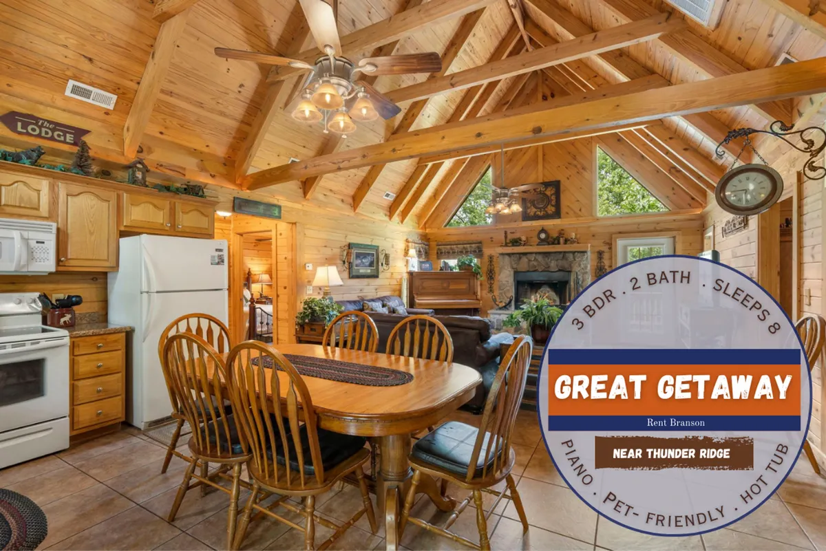 A warmly lit interior scene featuring a spacious dining table, exposed wooden beams, a stone fireplace, and a classic piano in the background. The arrangement invites family gatherings, meals, game nights, and musical evenings in a rustic cabin setting.