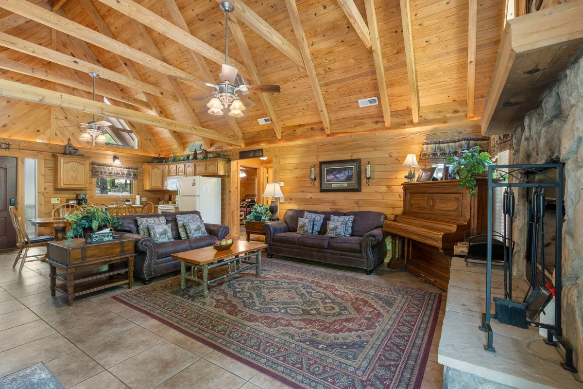 A cozy family room with wooden ceiling beams, plush sofas, a decorative rug, a piano, and a roaring stone fireplace. The space looks welcoming and ideal for socializing, relaxing, and enjoying music by the fire.