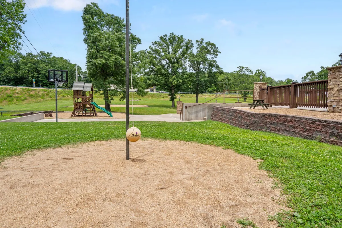 An exterior scene featuring a volleyball court, basketball hoop, playground equipment, and green grass. The area is set for active play, with evidence of sunny, communal gatherings.