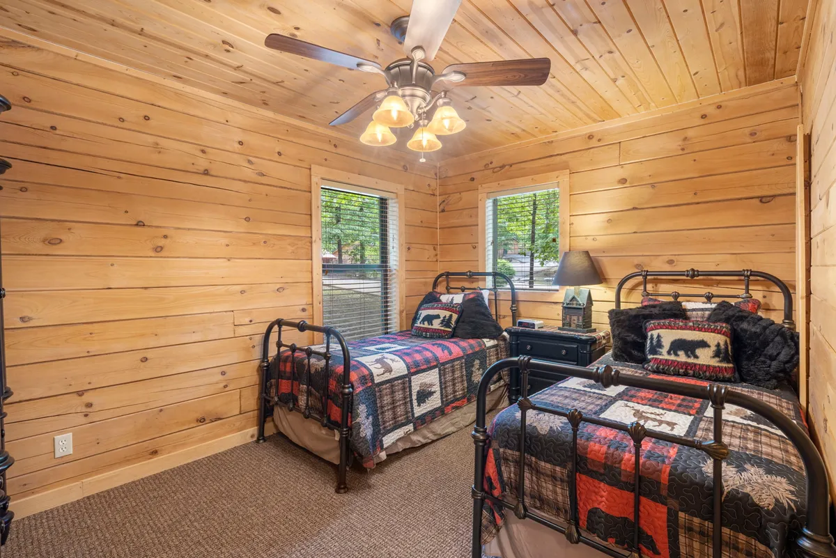 A double twin bedroom with metal bed frames, warm plaid bedspreads, wooden walls, and large windows letting in daylight. The beds are spaced for privacy and comfort, with a TV visible for entertainment.