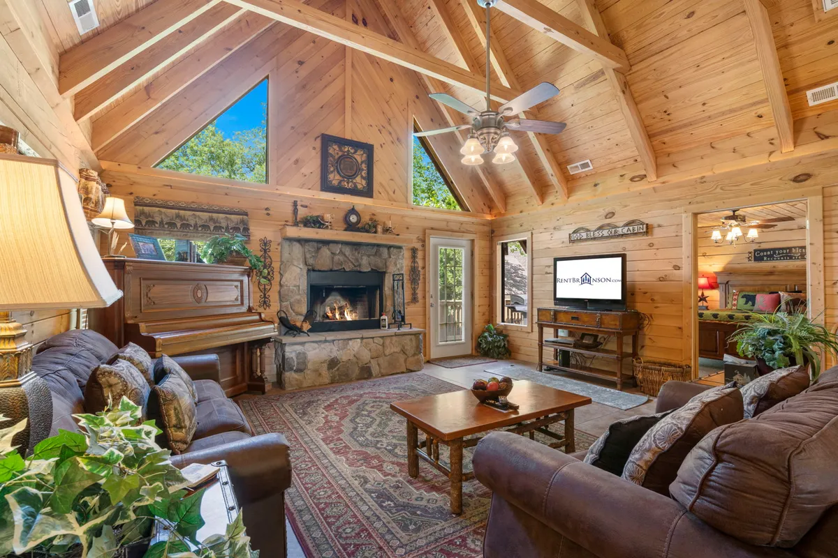 The interior of a spacious log cabin featuring soaring wooden ceilings, a striking stone fireplace at the heart of the room, large sunlit windows, and a piano inviting guests to create their own music. Cozy seating surrounds the hearth, making it perfect for family gatherings, relaxation, and memorable moments together.