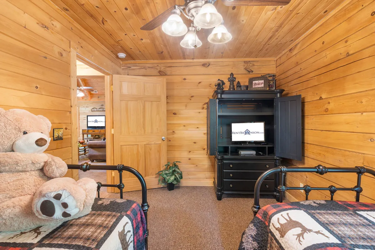 A welcoming twin bedroom with wooden paneling, twin beds dressed in cozy checkered blankets, a large teddy bear, and a black TV cabinet. The arrangement is both playful and inviting, ideal for families or friends.