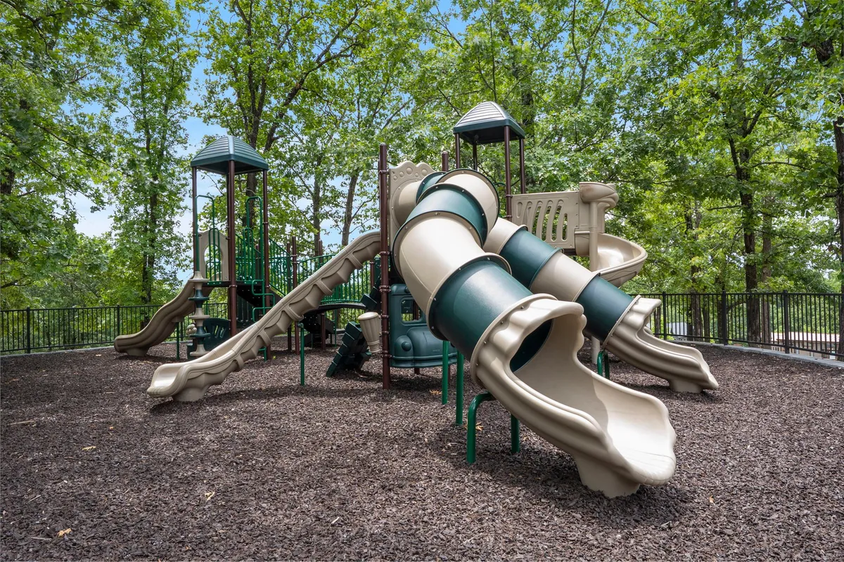 A lively exterior shot of the playground area, featuring playground slides, climbing structures, and mature trees. Children are seen playing, and families are enjoying the outdoors, which adds vibrancy and energy to the scene.