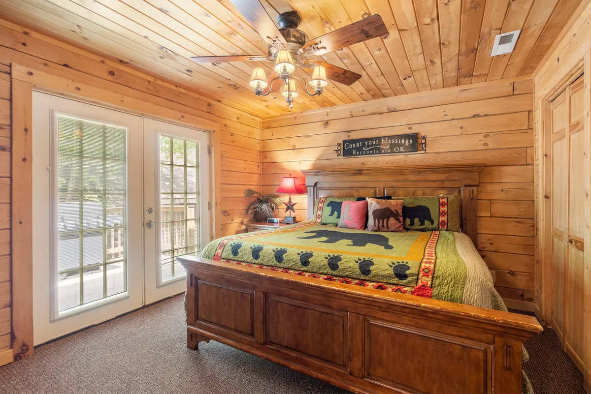 A cozy bedroom featuring a sturdy wooden bed frame with colorful bedding, sliding glass doors that invite natural light, and a ceiling fan overhead. The rustic decor and wood finishes evoke an authentic cabin feel, while a television sits across from the bed for personalized relaxation.