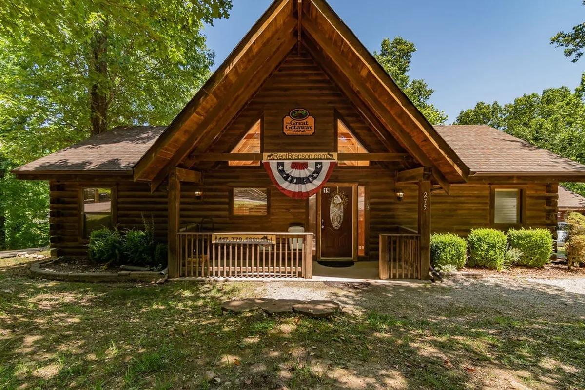 The image shows a charming log cabin exterior with a welcoming front porch and an American flag banner. The cabin is surrounded by nature, highlighting the seamless connection between the accommodation and the outdoors.