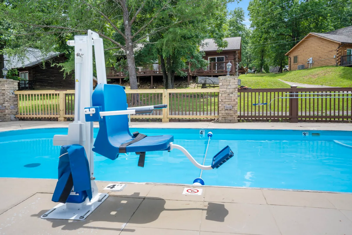 This exterior image captures the resort's swimming pool with an ADA pool lift visible, surrounded by lounge chairs, trees, and cottages in the background. The scene shows a welcoming, accessible pool area perfect for all guests.